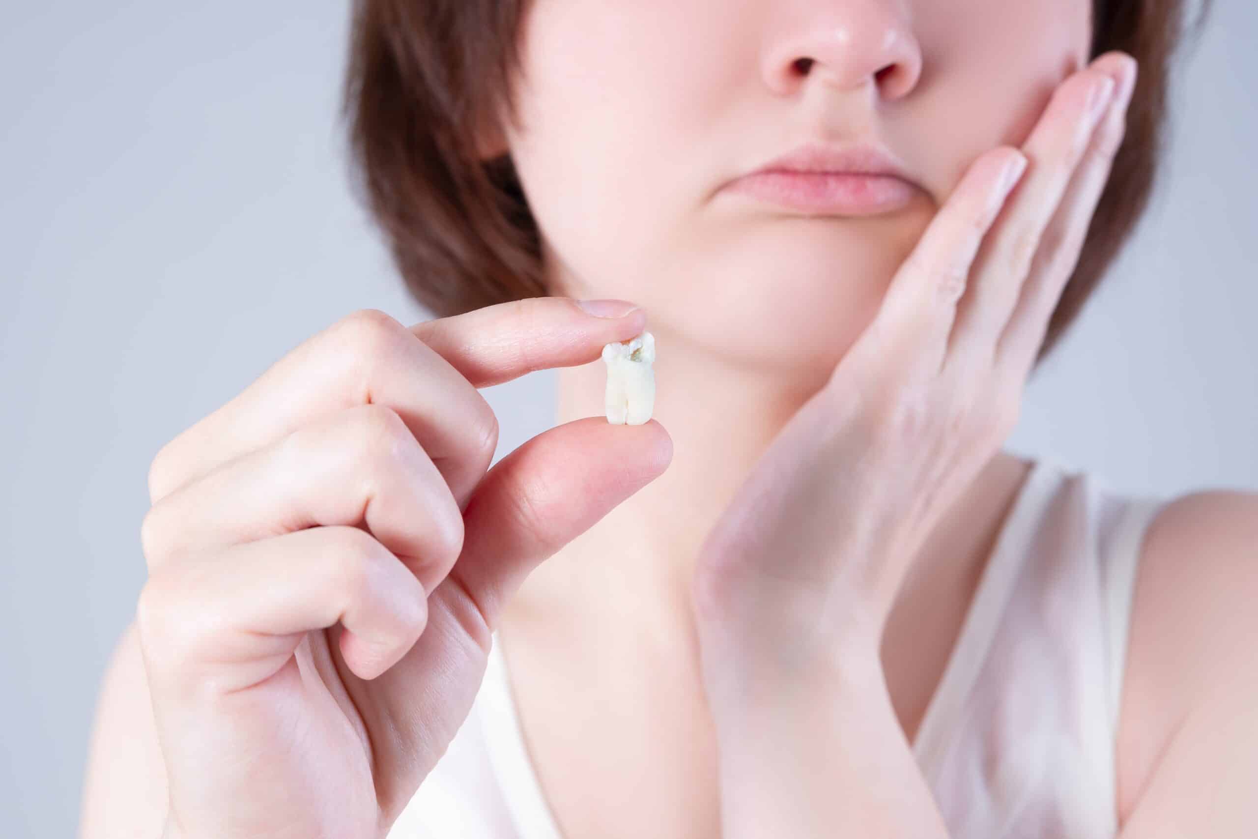 A woman visible from the nose down holds out a tooth with one hand and holds her jaw with the other, frowning to indicate pain.